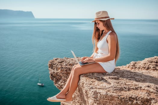 Successful business woman in yellow hat working on laptop by the sea. Pretty lady typing on computer at summer day outdoors. Freelance, travel and holidays concept.