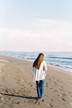 Young woman in a long sweater walks along the sandy beach. Back view. High quality photo