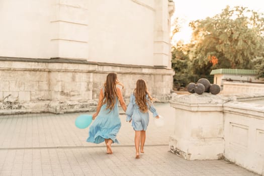 Daughter mother run holding hands. In blue dresses with flowing long hair, they hold balloons in their hands against the backdrop of a sunset and a white building