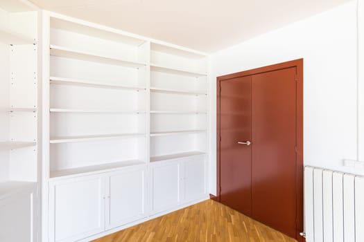 Large wooden closed mahogany door with a brass handle for opening. Along the wall is a white open cabinet with shelves for books and interior items for coziness and comfort. The floor is parquet