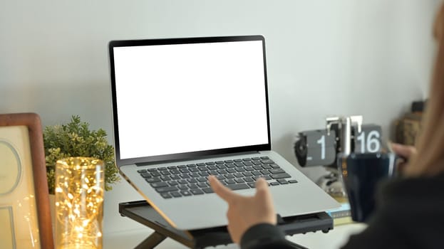 Cropped view of young woman holding cup of coffee and pointing at blank laptop computer screen.