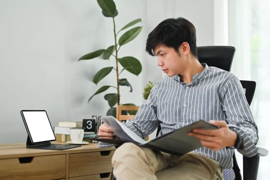 Handsome asian man freelancer looking at digital tablet screen while working remotely from home.