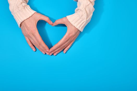 Top view of anonymous person in sweater showing heart shaped gesture with hands against blue background