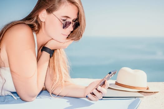 Successful business woman in yellow hat working on laptop by the sea. Pretty lady typing on computer at summer day outdoors. Freelance, travel and holidays concept.