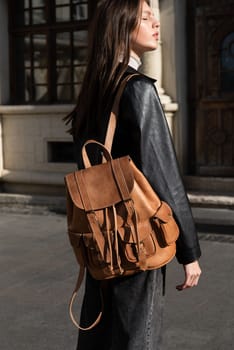 beautiful young girl with dark hair wearing jeans and a leather jacket posing outside with a leather backpack