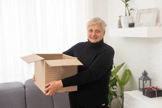 Smiling elderly woman customer receive post shipment parcel at home, happy old senior grandma hold open cardboard box sit on sofa in living room, online shopping order fast courier delivery concept