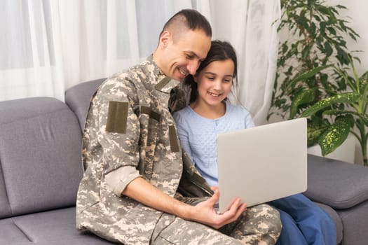 Smiling father in camouflage uniform near daughter, laptop at home.