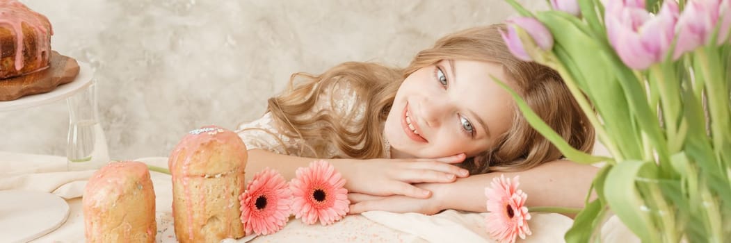 A girl with long hair in a light dress is sitting at the Easter table with cakes, spring flowers and quail eggs. Happy Easter celebration