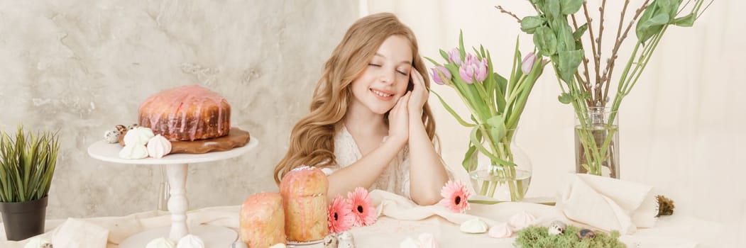 A girl with long hair in a light dress is sitting at the Easter table with cakes, spring flowers and quail eggs. Happy Easter celebration