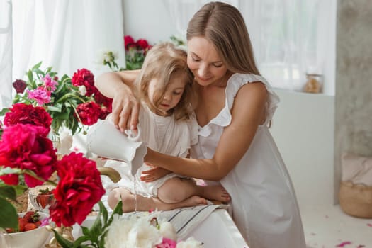 A little blonde girl with her mom on a kitchen countertop decorated with peonies. The concept of the relationship between mother and daughter. Spring atmosphere.