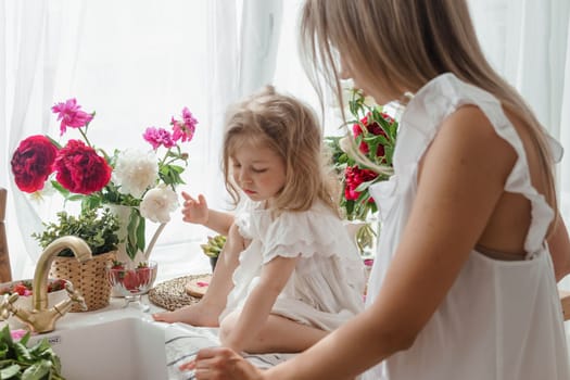 A little blonde girl with her mom on a kitchen countertop decorated with peonies. The concept of the relationship between mother and daughter. Spring atmosphere.