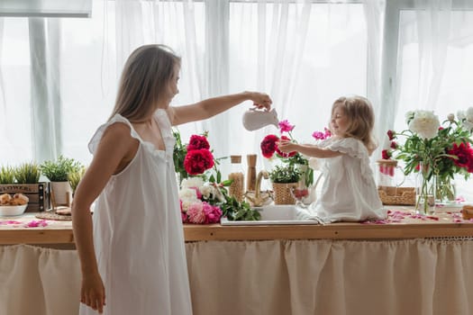 A little blonde girl with her mom on a kitchen countertop decorated with peonies. The concept of the relationship between mother and daughter. Spring atmosphere.