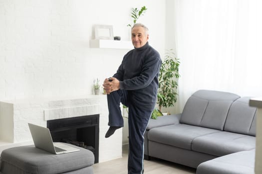 Sport in mature age. Happy middle-aged man doing stretching exercises in front of laptop at home, watching online tutorials, in living room interior, free space.