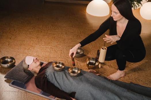 A young beautiful woman is doing a massage with singing bowls and a koshi bell to another girl. Sound therapy.