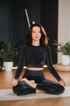 A woman in a black tracksuit sits on a yoga mat in a lotus position in the gym.