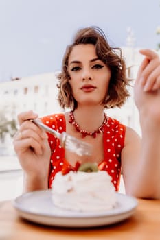 Charming woman in a restaurant, cafe on the street. She sits at the table and eats a cake with a fork. Dressed in a red sundress with white polka dots