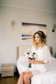 Bride in a white bathrobe sits with a bouquet of flowers on the bed in the room. High quality photo