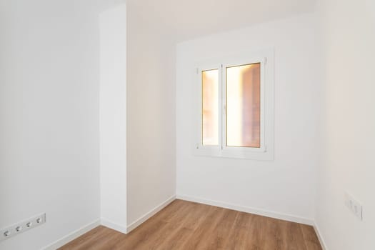 Part of an empty room with white walls, brown parquet. Plastic window in wall with frosted translucent glass. Socket is built into wall for simultaneous connection of several electrical appliances