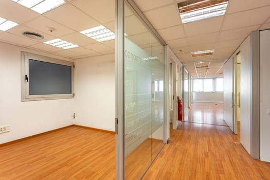 Glass partition enclosing empty office room from corridor with window at end. Outdated design of office corridor and room that needs to be renovated. Burnt out fluorescent lamps in empty office space