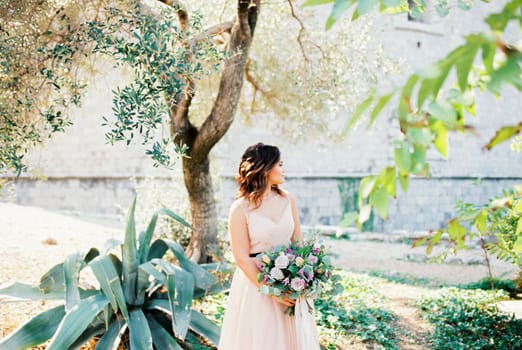 Bride with a bouquet stands near a green agave bush in front of a stone fortress and looks away. High quality photo