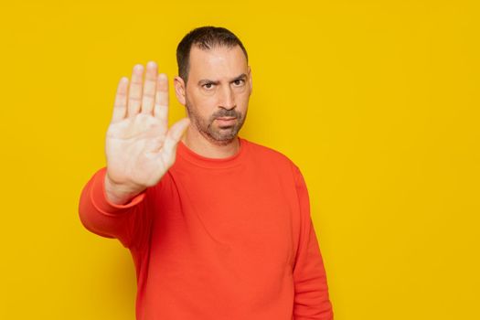 Hispanic man with beard in red sweater making stop gesture while looking angrily at camera, isolated on yellow background