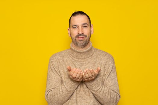 Hispanic man with a beard wearing a beige turtleneck advertising something on his hands, isolated over yellow background