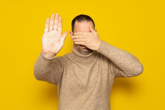 Man in his 40s with a beard wearing a turtleneck covering his eyes with his hands and making a stop gesture with an expression of sadness and fear. Embarrassed and negative concept.