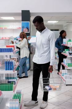 African american man shopping in drugstore, buying medications, reading instruction on tablets package, standing in pharmacy aisle. Client choosing vitamins, purchasing medicaments, wide shot