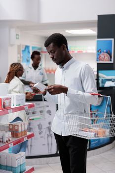 African american customer shopping in drugstore, buying supplements, reading instruction on tablets package. Young man choosing vitamins, purchasing medicaments in pharmacy store, medium shot