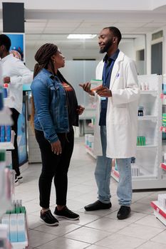 African american man pharmacist recommending woman vitamin c in drugstore. Nutritional supplements retail, client and pharmacy store consultant talking, buyer choosing pills, wide shot
