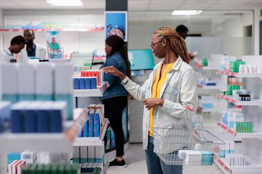 African american woman checking medications in drugstore shelf, looking at tablet packages. Client choosing nutritional supplements, customer standing in pharmacy aisle, medium shot