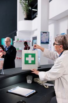 Elderly pharmacist standing at drugstore counter putting pills packages in paper bag while waiting for client to come and pay for prescription treatment. Health care service and support