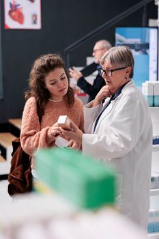 Drugstore worker checking box of pills explaining medical leaflet to caucasian client while working in pharmacy. Diverse customers looking at shop shelves shopping for pharmaceutics treatment