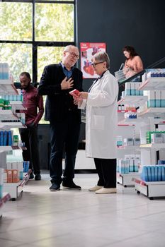 Elderly client asking for pharmaceutical treatment advice to drugstore employee in pharmacy, discussing cardiology pills leaflet. Customer doing health care shopping buying medication, vitamins