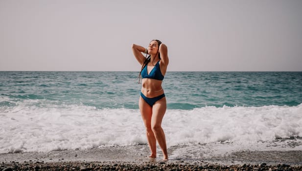 Beach vacation. Hot beautiful woman in sunhat and bikini standing with her arms raised to her head enjoying looking view of beach ocean on hot summer day.