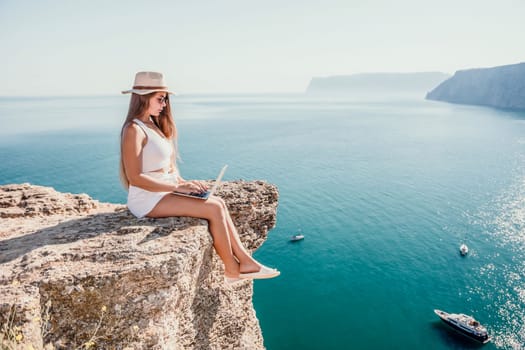 Successful business woman in yellow hat working on laptop by the sea. Pretty lady typing on computer at summer day outdoors. Freelance, travel and holidays concept.