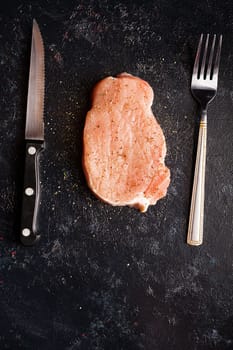 Raw steak meat on black wooden table next to fork and knife. Seasoned piece of meat. Gourmet food and fresh uncooked meal