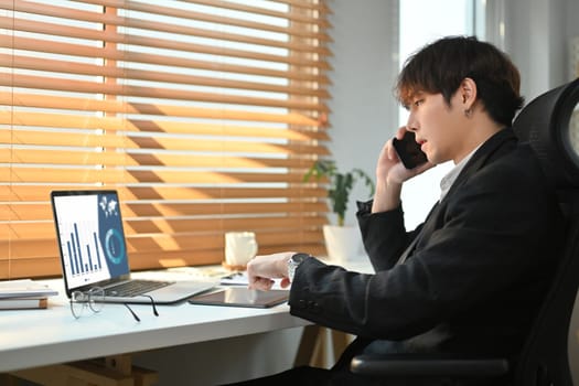 Millennial male business executive looking at laptop computer screen, discussing project on mobile phone.