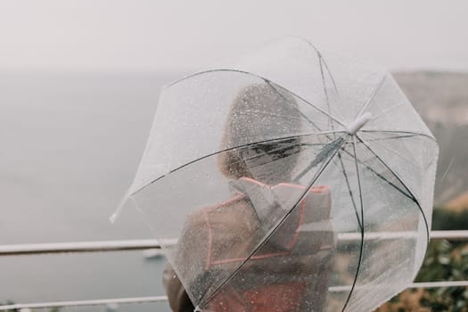 Woman rain park. Happy woman portrait wearing a raincoat with transparent umbrella outdoors on rainy day in park near sea. Girl on the nature on rainy overcast day