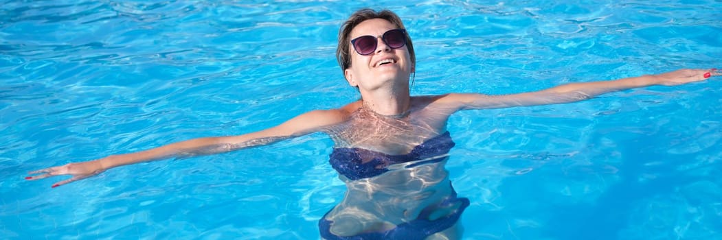 Portrait of smiling girl in sunglasses in swimsuit swims in pool. Pleasure and relaxation in summer pool