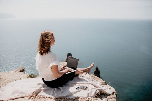 Successful business woman in yellow hat working on laptop by the sea. Pretty lady typing on computer at summer day outdoors. Freelance, travel and holidays concept.