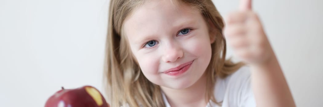 Cute little girl holds apple in one hand and thumbs up. Benefits of apples and fruits for children concept