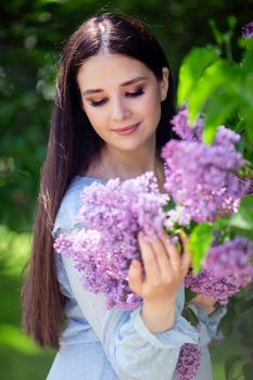 A happy brunette with long hair stands with lilac flowers, in the spring in the park, on a sunny day. Vertical. Close up