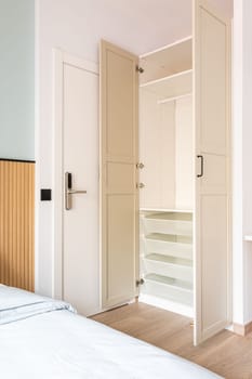Built-in wooden wardrobe with open doors in beige. Inside the cabinet, all shelves, drawers, crossbars are made of white durable plastic. Entrance to the hotel room with an electronic lock