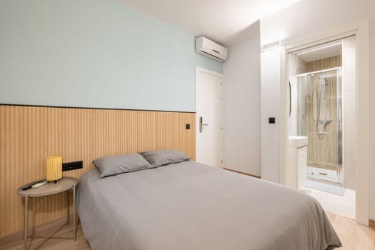 Bedroom with spacious bed against wall demarcated with wood paneling and blue paint and bedside table. Bedding is in beautiful gray color. From room there is doorway to bathroom with glass shower