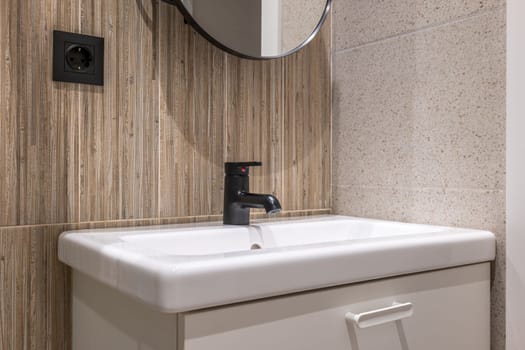 Bathroom sink on vanity is illuminated by bright light from ceiling. Walls are combination of gray granite and wood grain marble tiles. Above sink is mirror in black frame, like faucet and an outlet