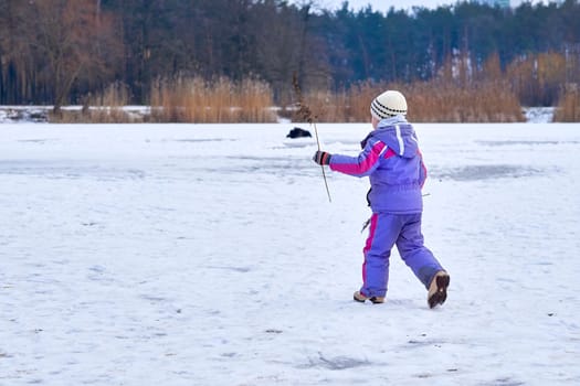 a young human being below the age of puberty or below the legal age of majority. A cute child in a lilac jumpsuit walks on the lake in a winter park.