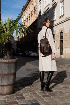 beautiful young girl with dark hair wearing leggings and a beige coat posing outside with a leather backpack