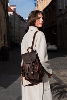 beautiful young girl with dark hair wearing leggings and a beige coat posing outside with a leather backpack