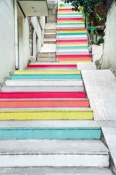 Stairs are painted with multicolored paint, rising up narrow street, view from below, concept of positivity